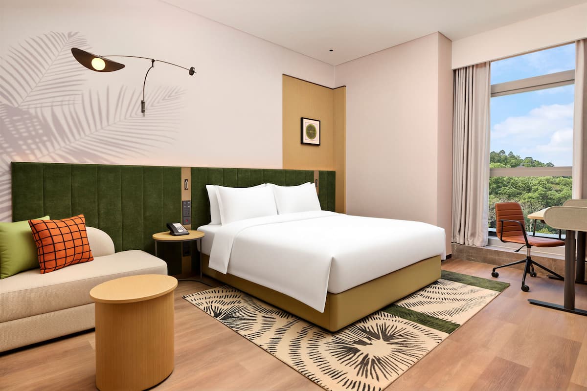 A hotel room with a bed, a sofa, a desk, a chair, a lamp, and a window with a view.