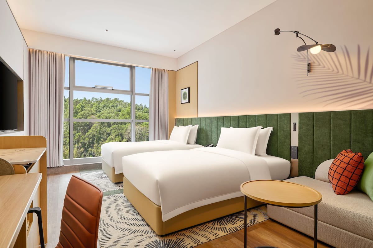 A hotel room with two beds, a sofa, a small table, a TV, and a large window with a view of trees.