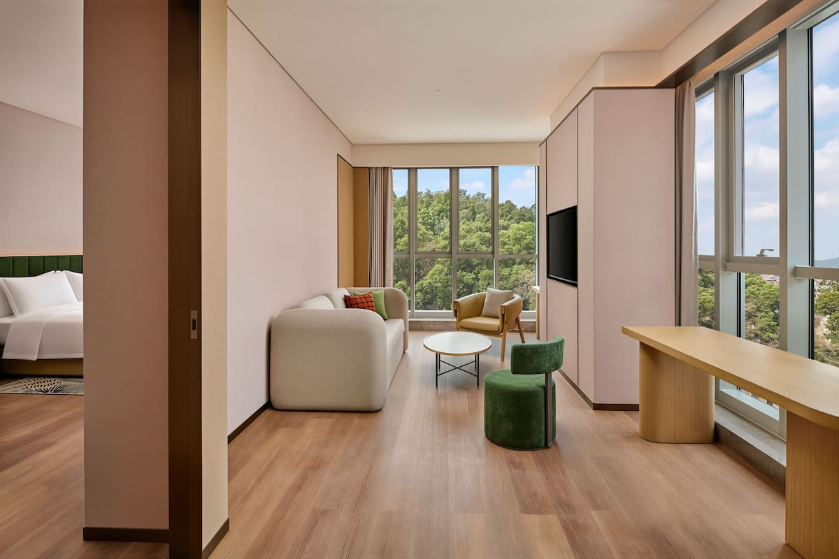 A modern hotel room with a large window, a sofa, a round coffee table, a green ottoman, and a wooden desk.