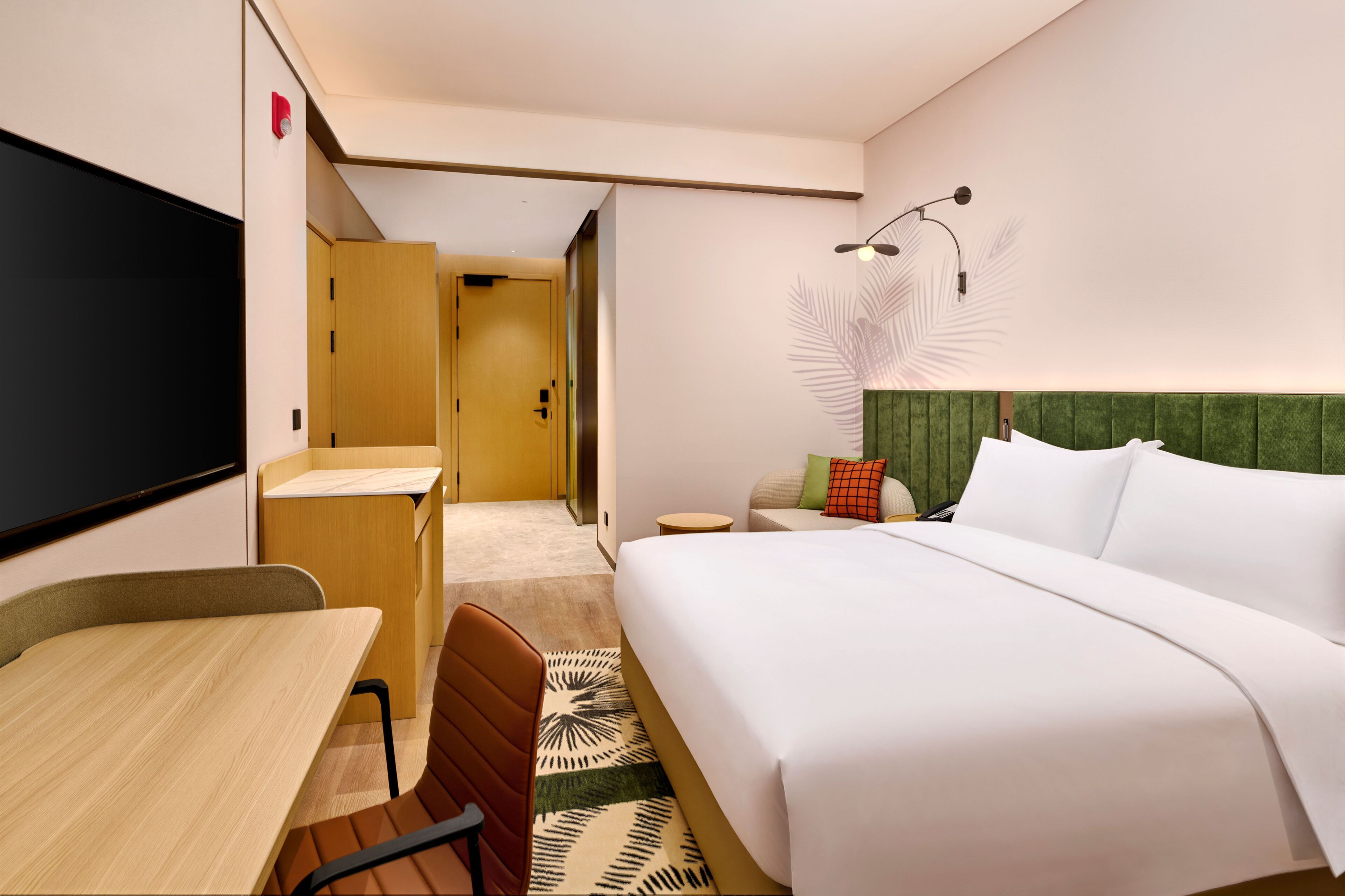 A hotel room with a bed, a desk, a chair, a TV, a bedside table, and a lamp.