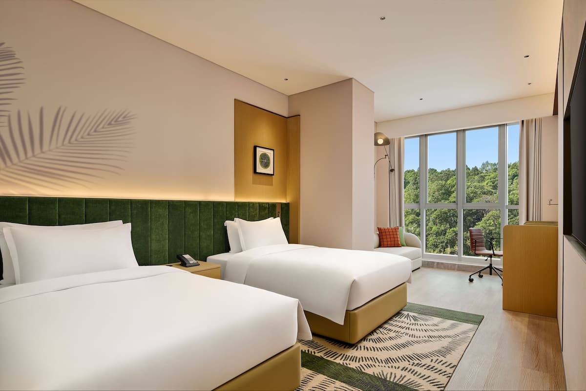 A hotel room with two beds, a desk, a chair, a large window with a view of trees, and a mounted television.
