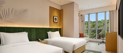 A hotel room with two beds, a desk, a chair, a large window with a view of trees, and a mounted television.