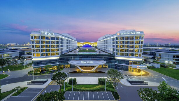 A modern hotel with a central atrium, two wings labeled 'Hilton' and 'Hilton Garden Inn,' and a parking area in front. - Hilton Garden Inn Guangzhou Baiyun Airport Terminal 3 (Guangzhou)
