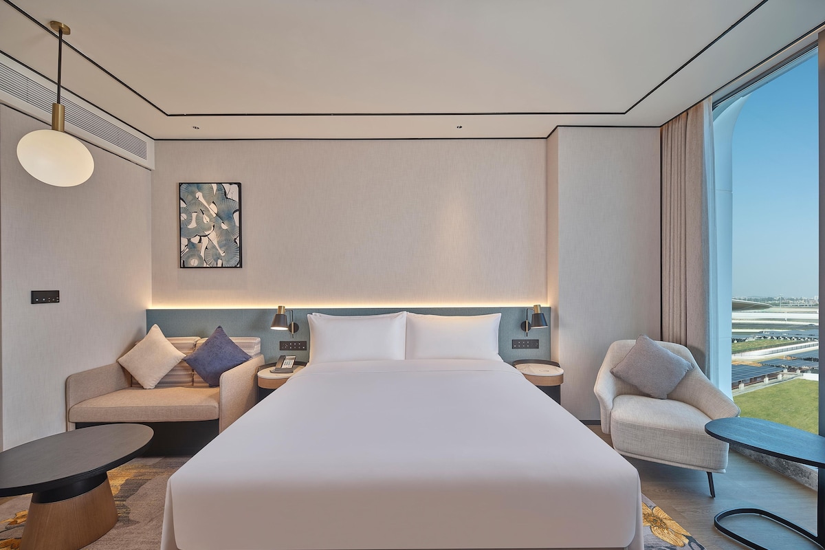 Una habitación de hotel moderna con una cama grande, un sofá, una silla, una mesita y vistas al aeropuerto.