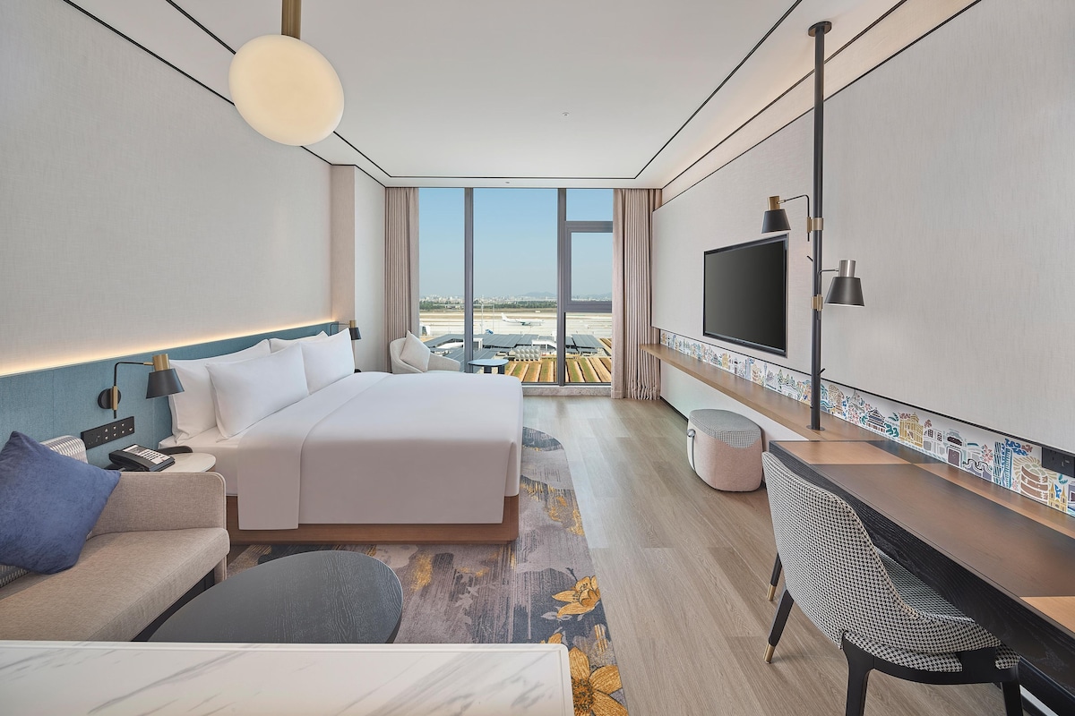 Ein modernes Hotelzimmer mit einem großen Bett, einer Sitzecke, einem Schreibtisch, einem Fernseher und Blick auf einen Flughafen.