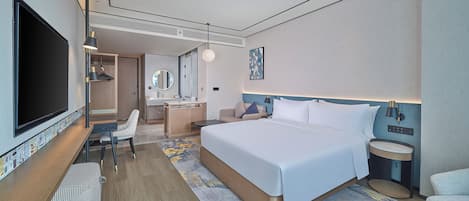 A modern hotel room with a large bed, a desk, a chair, a TV, and a bathroom with a mirror and sink.