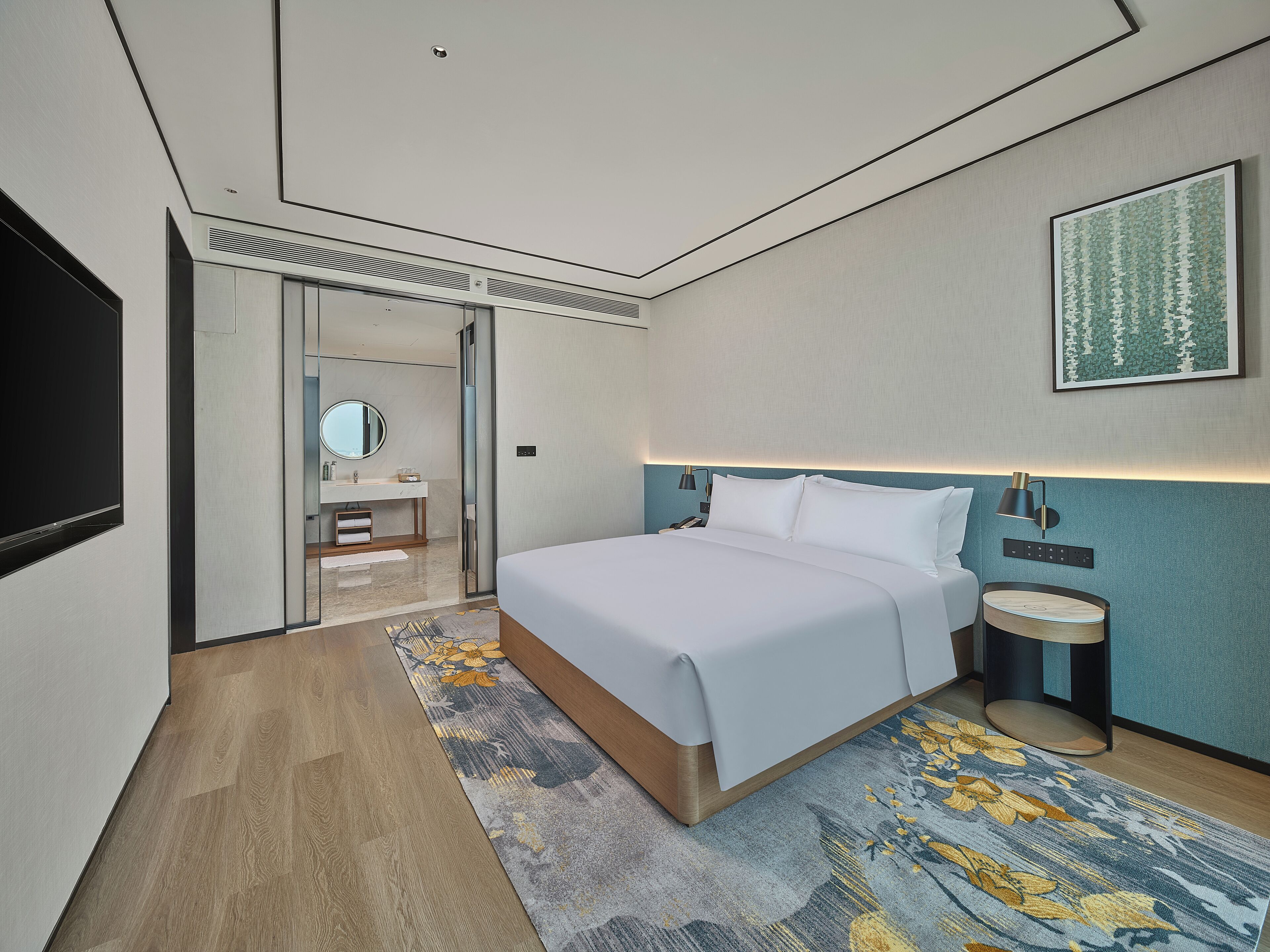 A modern hotel room with a large bed, a bedside table with a lamp, a small round table, and a mirror.