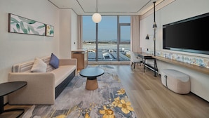 A modern hotel room with a large window offering a view of an airport, a flat-screen TV, a sofa, a coffee table, and a desk with a chair. - Hilton Garden Inn Guangzhou Baiyun Airport Terminal 3 (Guangzhou)