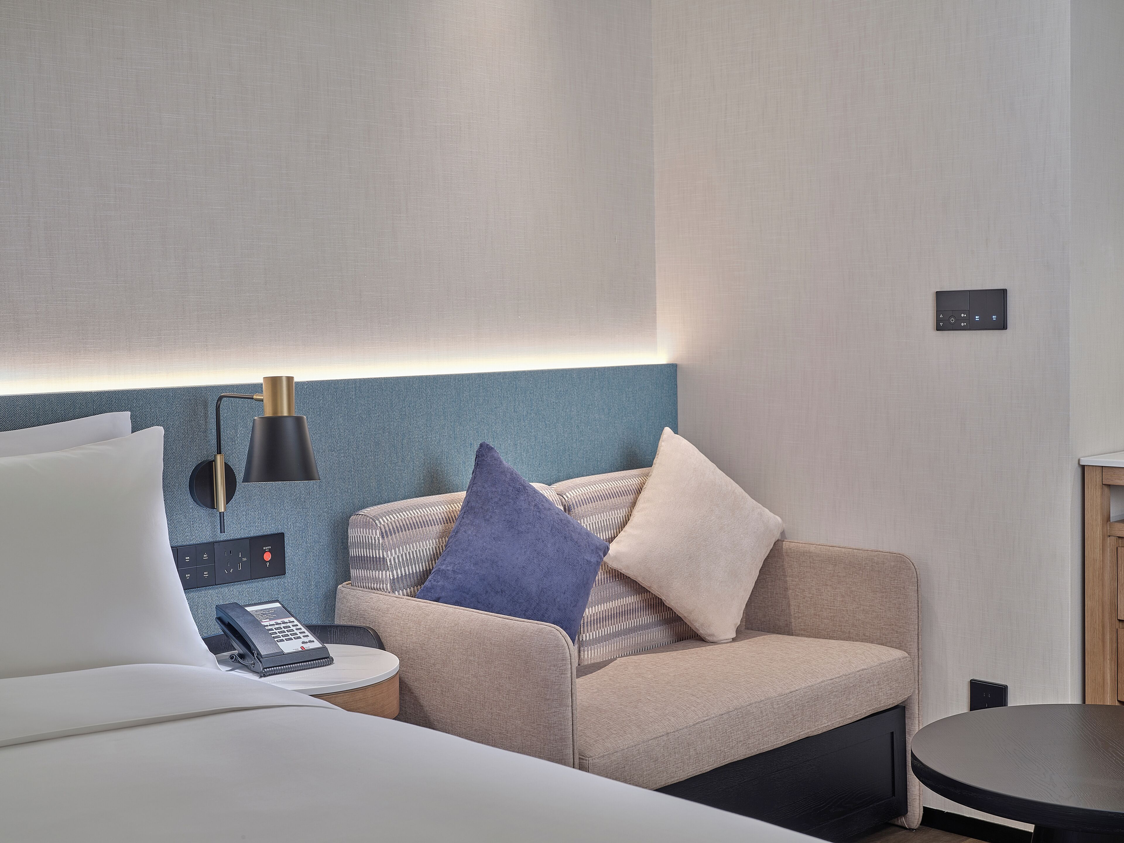 A modern hotel room with a bed, a small round table, a sofa with cushions, a lamp, and a control panel on the wall.