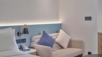 Una habitación de hotel moderna con una cama, una mesita redonda pequeña, un sofá con cojines, una lámpara y un panel de control en la pared.