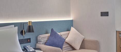 A modern hotel room with a bed, a small round table, a sofa with cushions, a lamp, and a control panel on the wall.