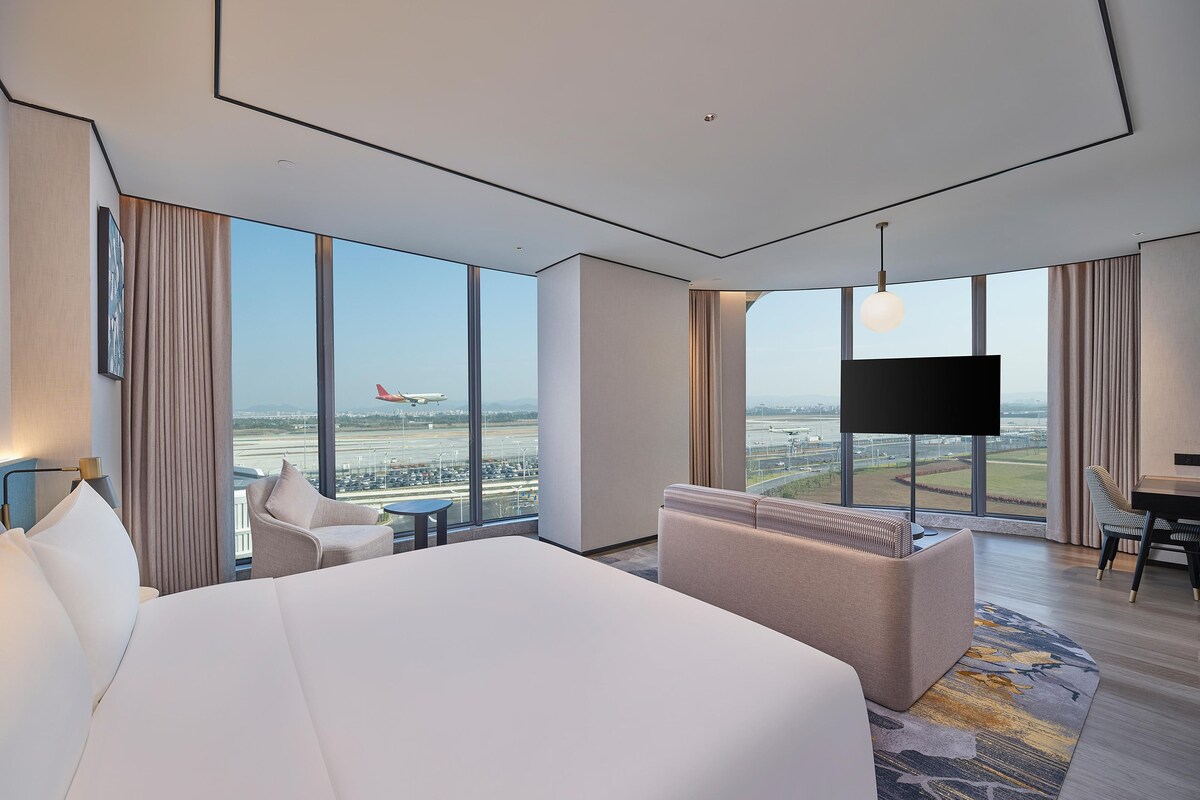 Ein Hotelzimmer mit einem großen Bett, einer Couch, einem Fernseher und Blick auf einen Flughafen.