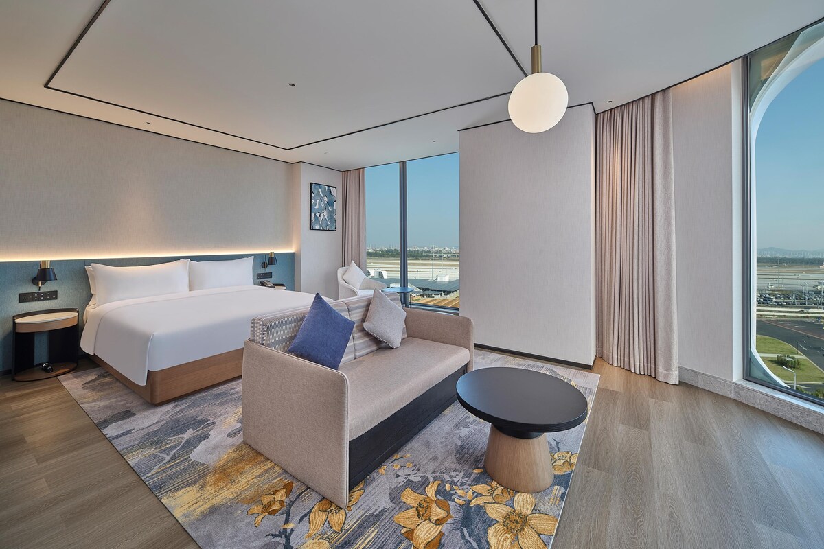 A modern hotel room with a large bed, a sofa, a small table, and a view of the airport.