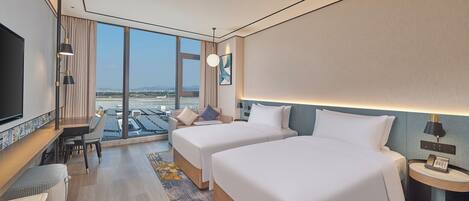 Une chambre d’hôtel moderne avec deux lits, un bureau et une grande fenêtre donnant sur un aéroport.