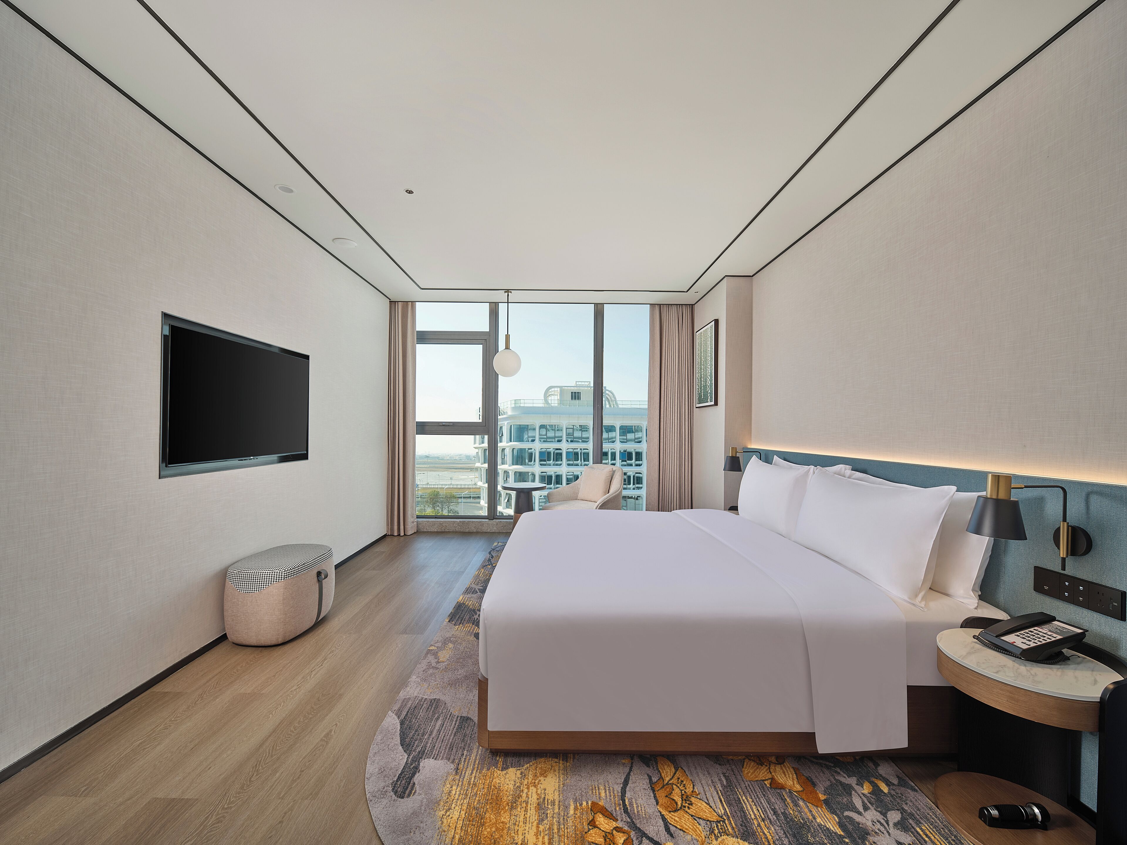 A modern hotel room with a large bed, a flat-screen TV mounted on the wall, a bedside table with a lamp, and a view of the city through large windows.
