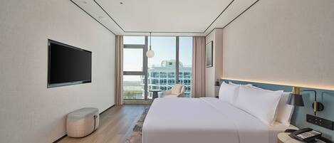 A modern hotel room with a large bed, a flat-screen TV mounted on the wall, a bedside table with a lamp, and a view of the city through large windows.