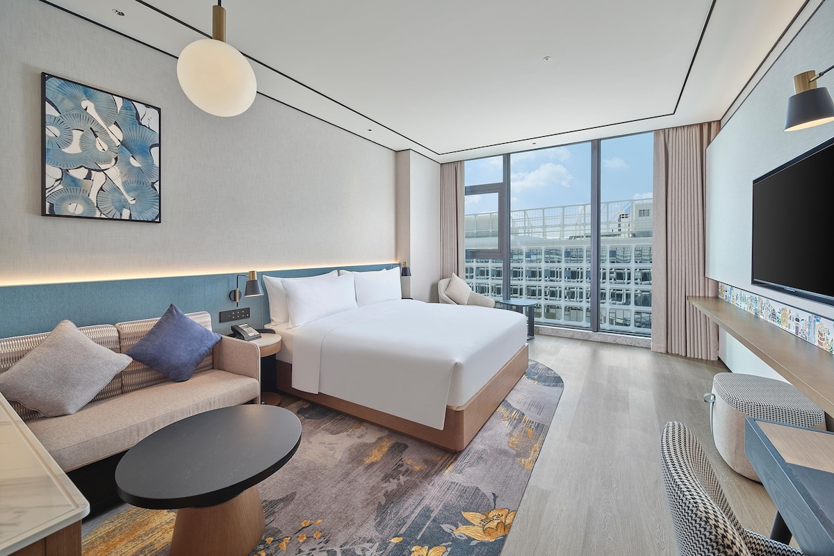 Una habitación de hotel moderna con una cama grande, un sofá, una mesita y una televisión, con vistas a la ciudad.