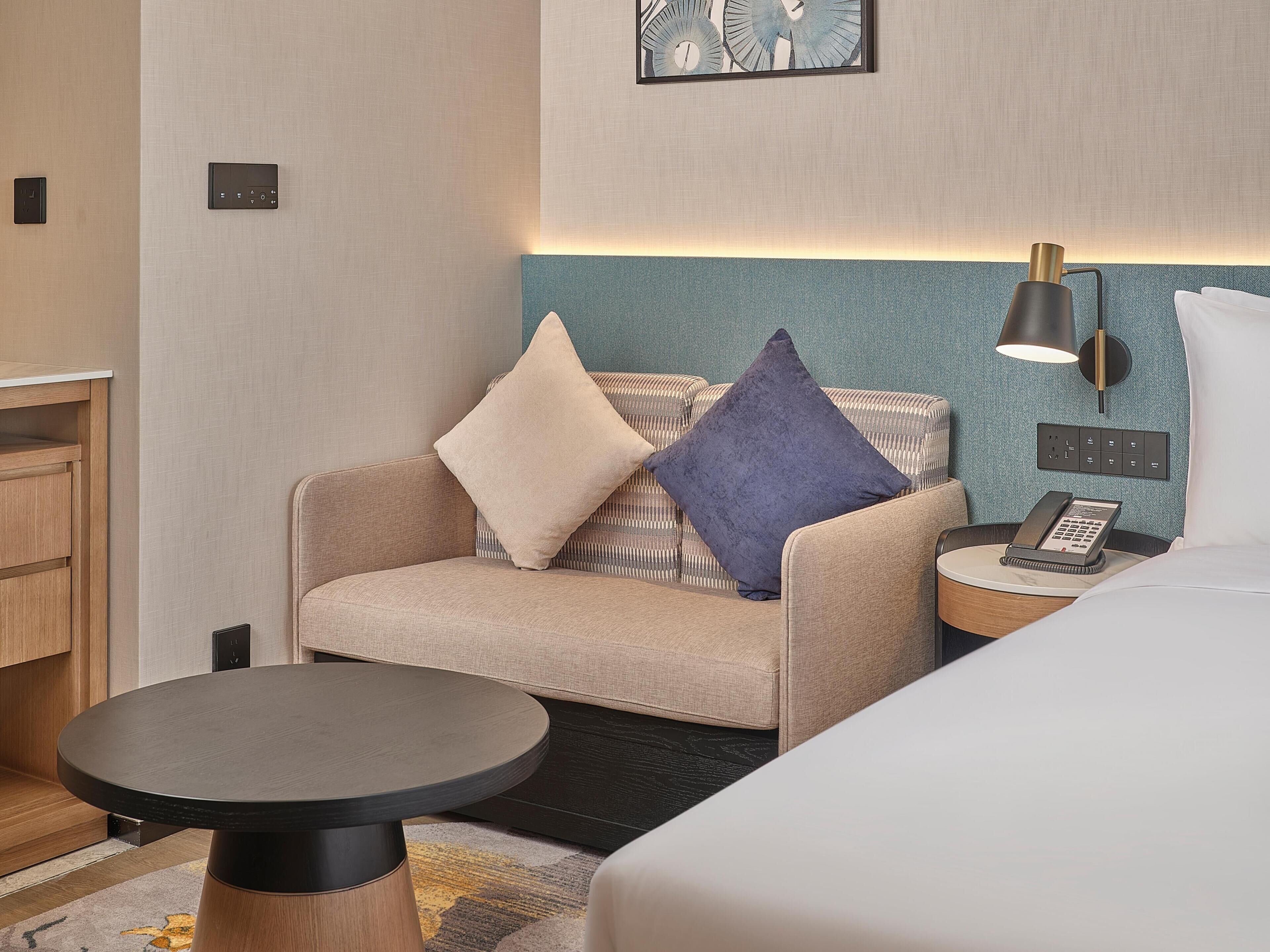 A hotel room with a sofa, a small table, a bedside table with a lamp, and a phone.