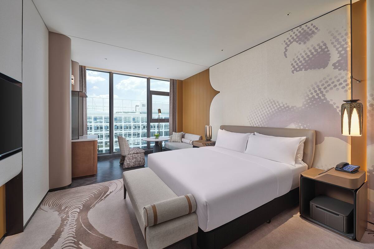 A modern hotel room with a large bed, a desk, and a view of the cityscape through large windows.
