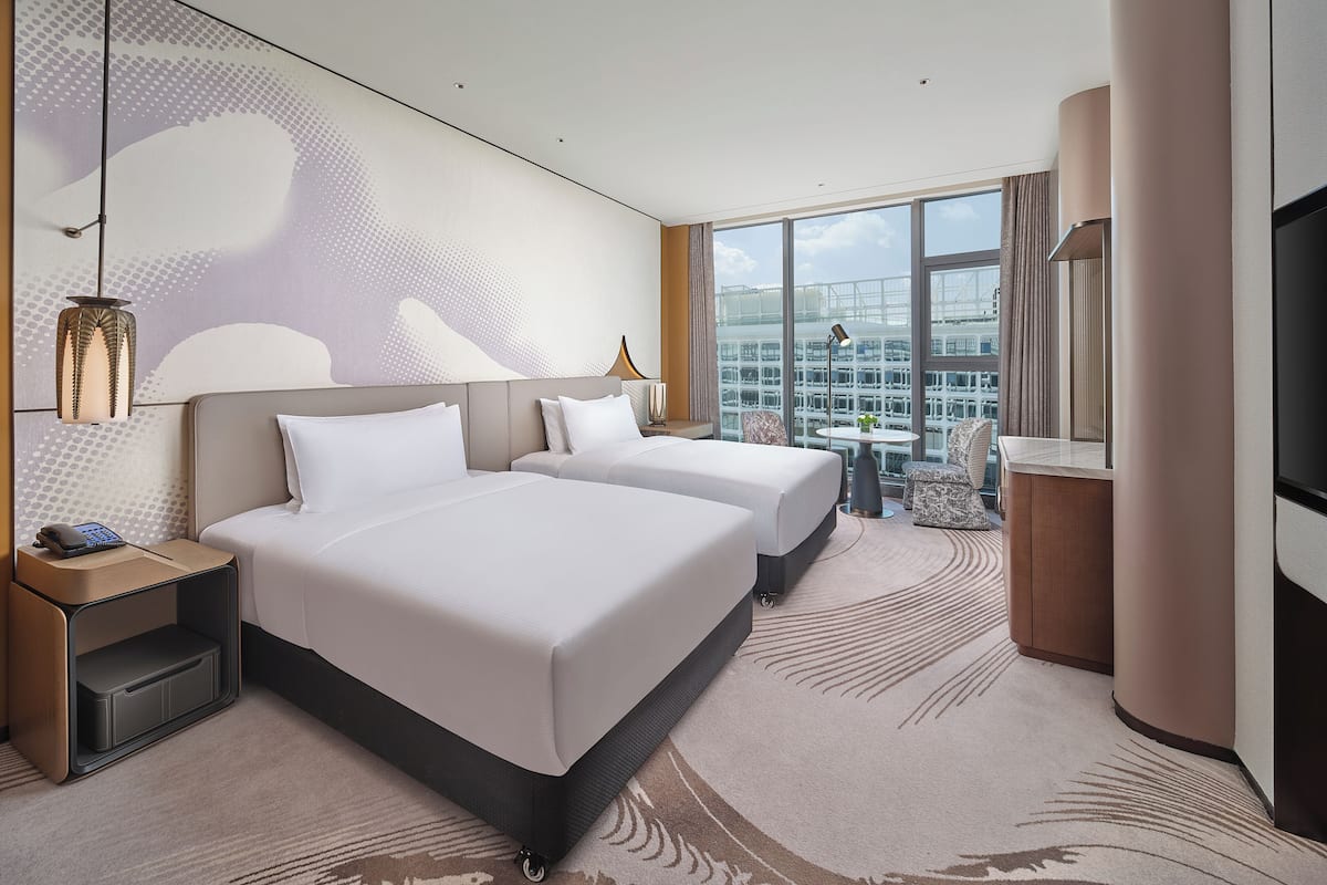 A hotel room with two beds, a desk, a chair, a TV, and a large window with a city view.