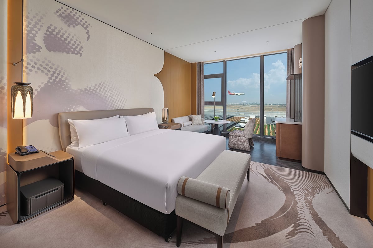 A modern hotel room with a large bed, a sofa, a small table, and a TV. There is a large window with a view of an airport.