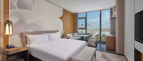 A modern hotel room with a large bed, a sofa, a small table, and a TV. There is a large window with a view of an airport.
