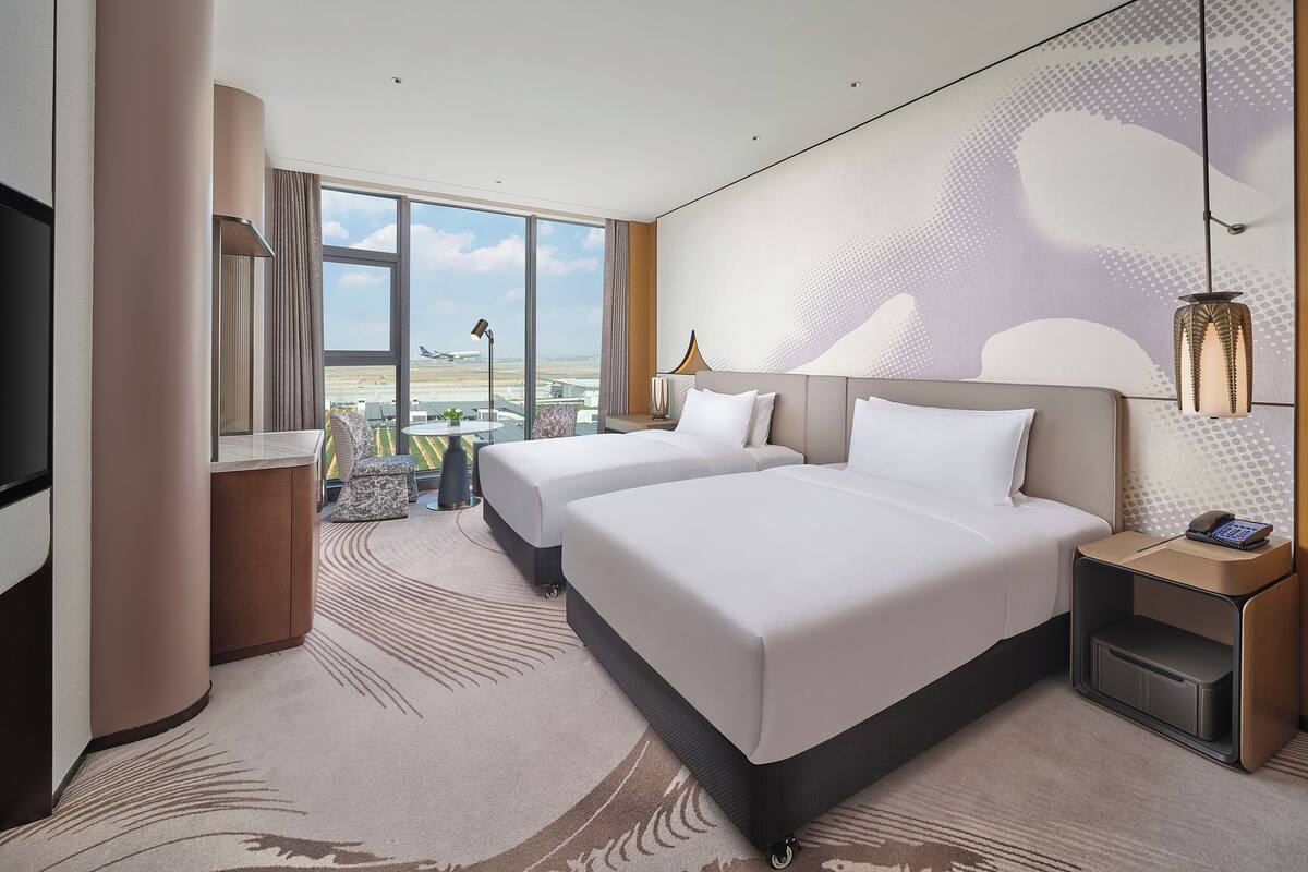 A hotel room with two beds, a TV, a desk, and a view of an airport.