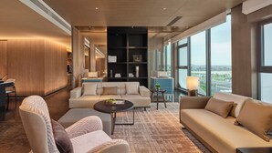 A modern hotel room with a large window, a sofa, a coffee table, and a bookshelf.