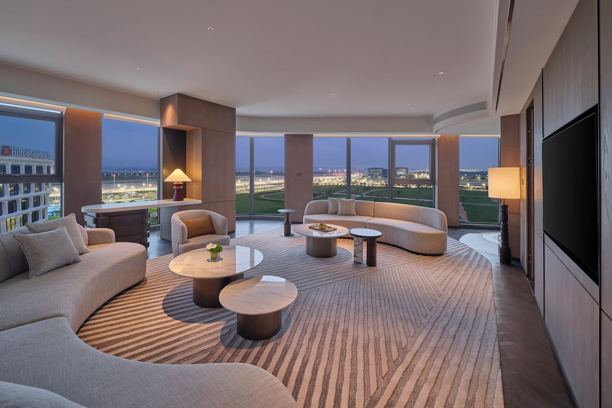 A modern hotel room with a large sofa, a smaller sofa, two round coffee tables, a TV, and a view of a cityscape.