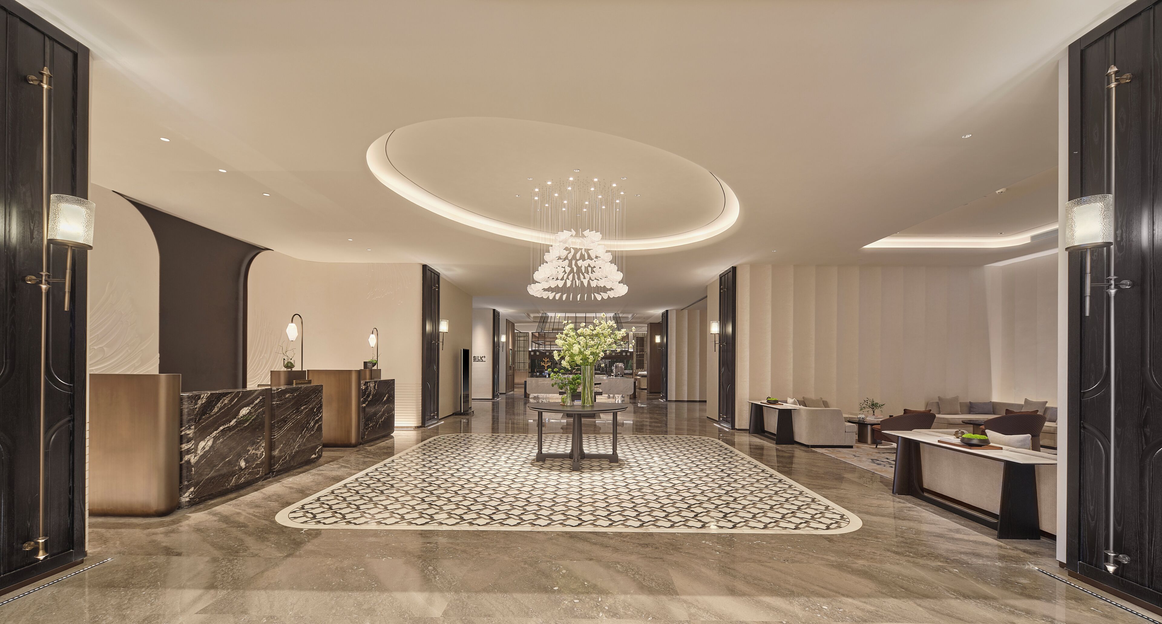 A modern hotel lobby with a marble reception desk, a central chandelier, and a patterned carpet.