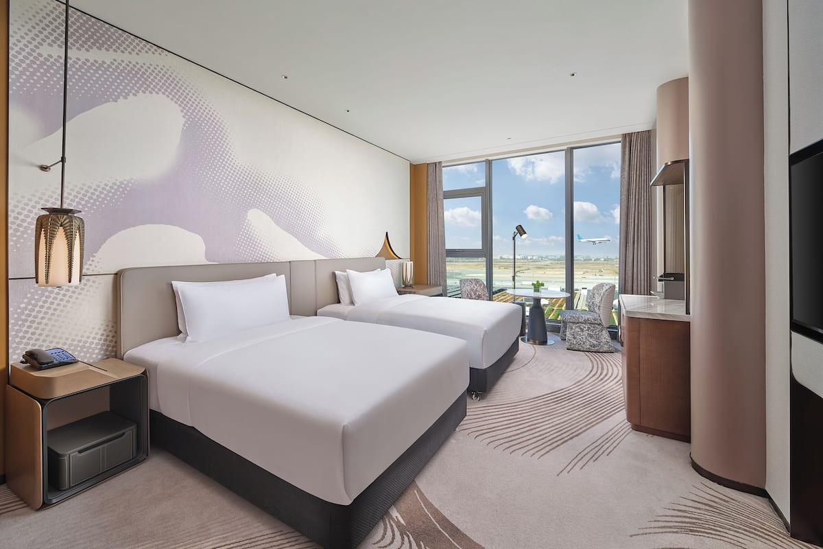 A hotel room with two beds, a large window with a view of the airport, a desk with a lamp, and a TV.