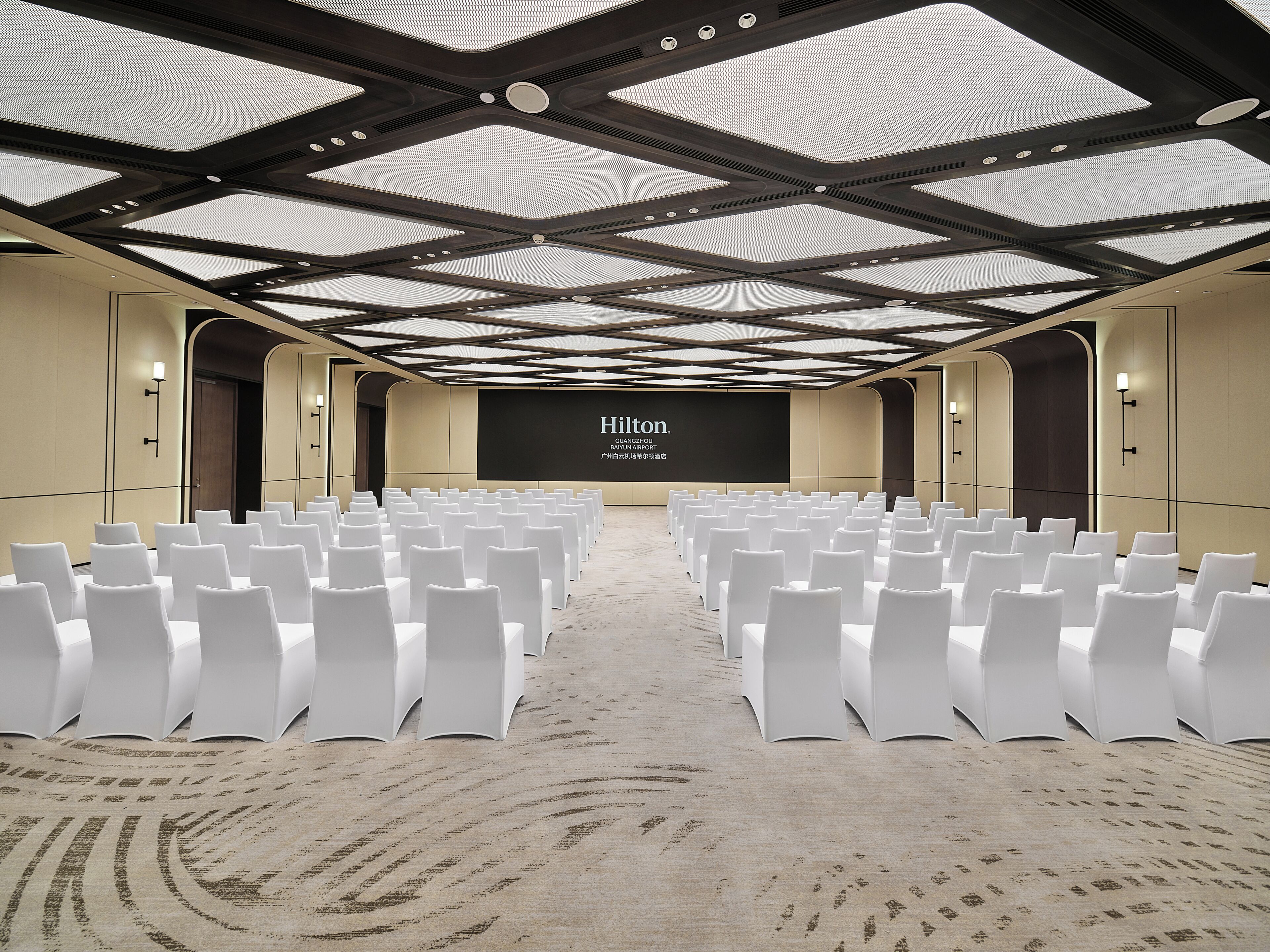 Une salle de conférence avec des rangées de chaises blanches, un grand écran et un design de plafond moderne.