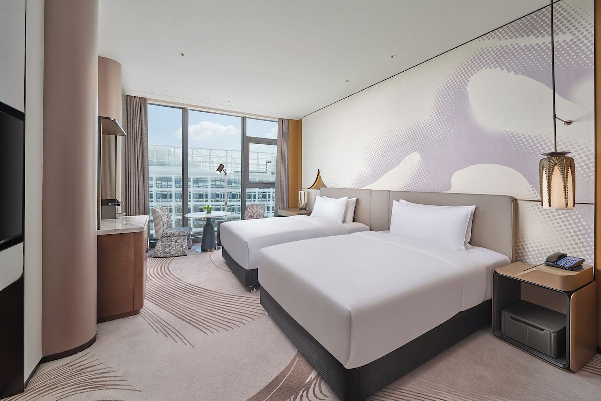A hotel room with two beds, a large window with a city view, a flat-screen TV, and a modern lamp.
