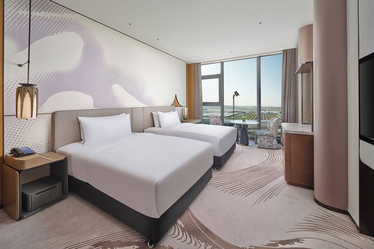 A hotel room with two beds, a large window, a TV, and a desk with a lamp.