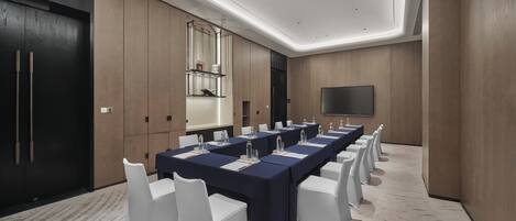 Un salón de conferencias con largas mesas, sillas blancas, un televisor de pantalla plana grande y un panel de pared de madera.
