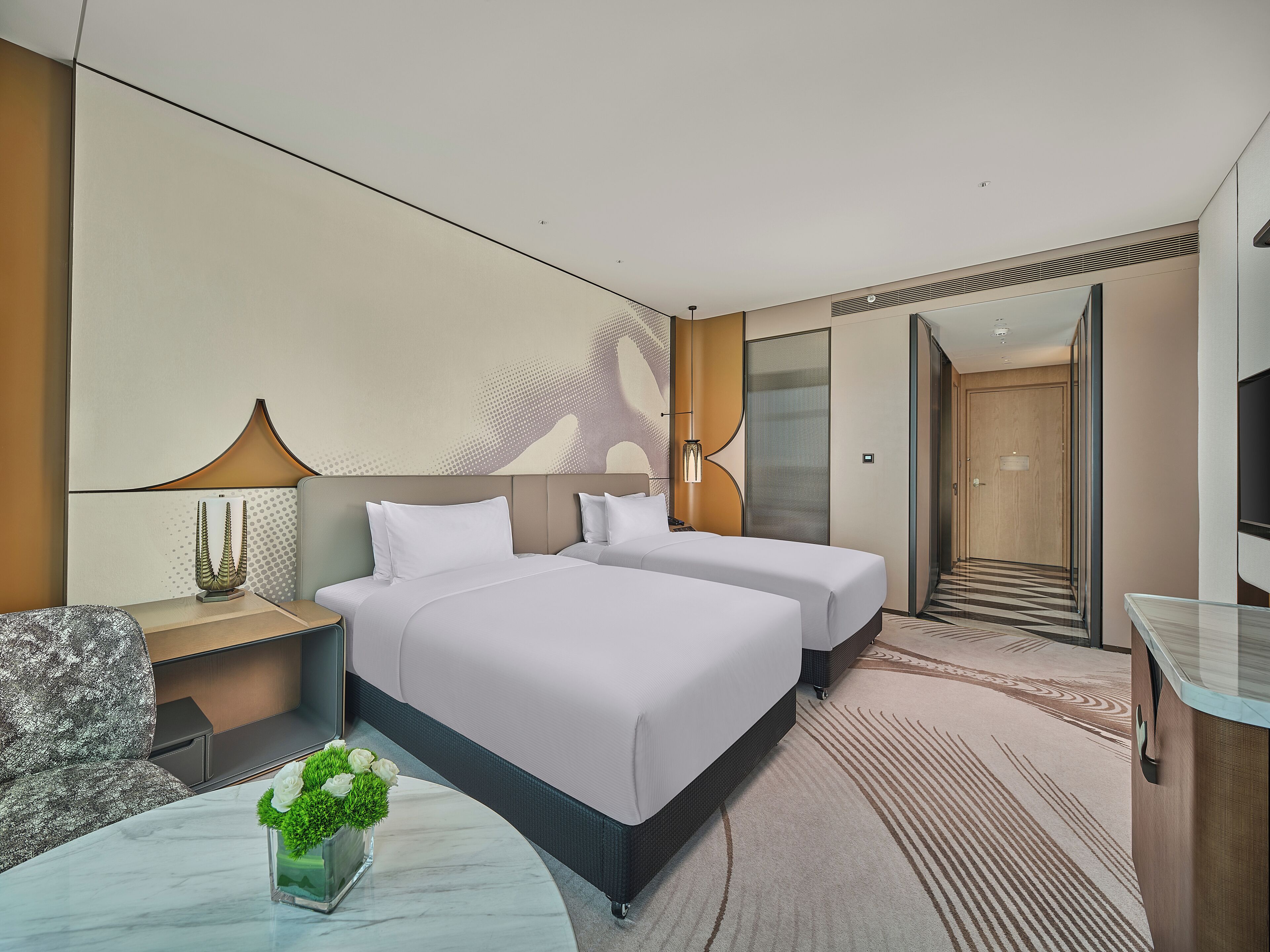 A modern hotel room with two beds, a sitting area with a chair, a small table with a vase of flowers, and a large mirror.