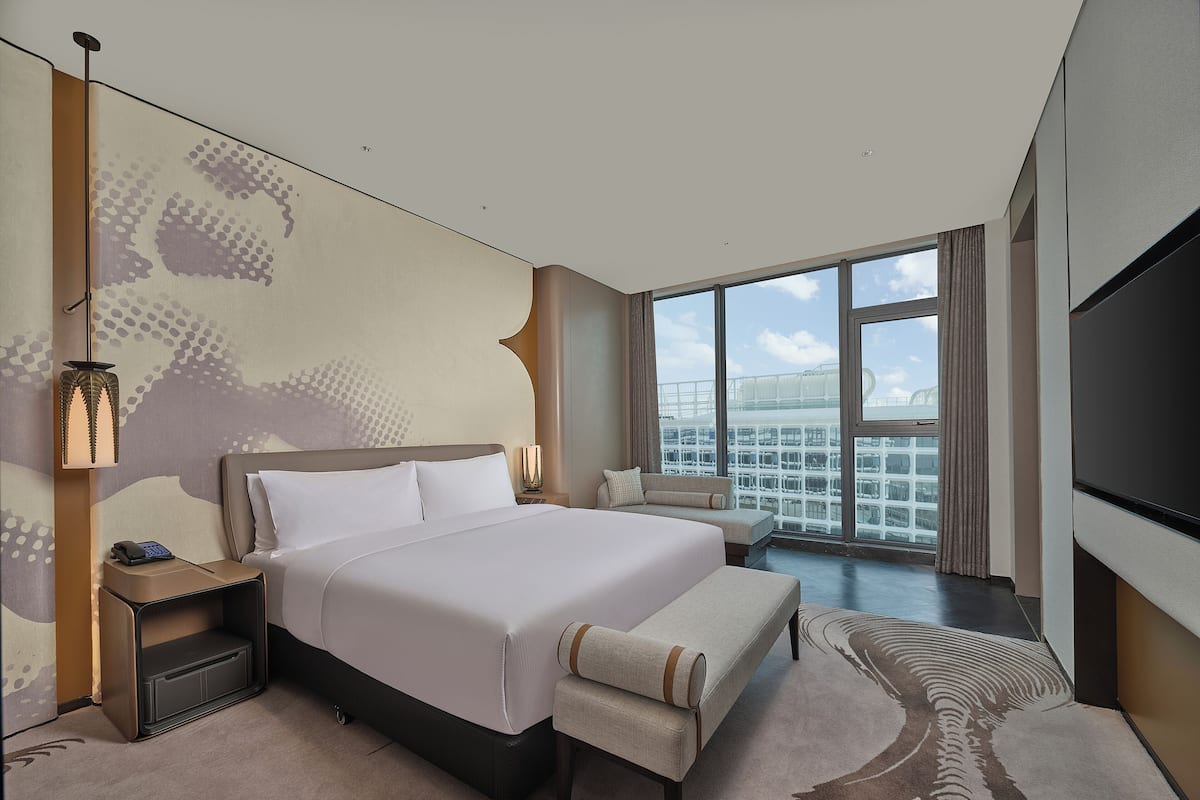 A modern hotel room with a large bed, a seating area, a TV, and a view of the cityscape.