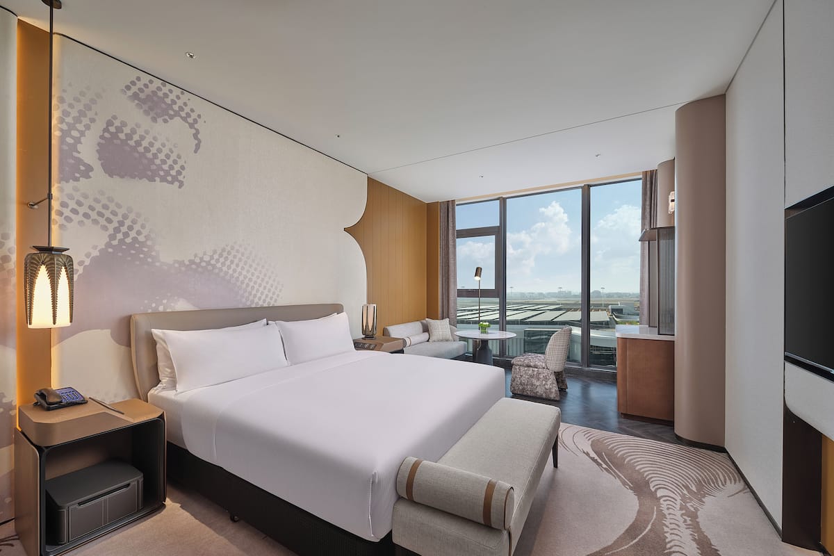 A modern hotel room with a large bed, a desk, a chair, a small table, and a TV. The room has a large window with a view of the city.