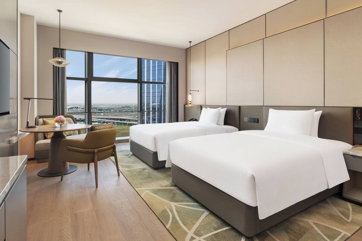 Ein modernes Hotelzimmer mit zwei Betten, einem Schreibtisch, einem Sessel, einem Fernseher und einem großen Fenster mit Blick auf die Stadt.
