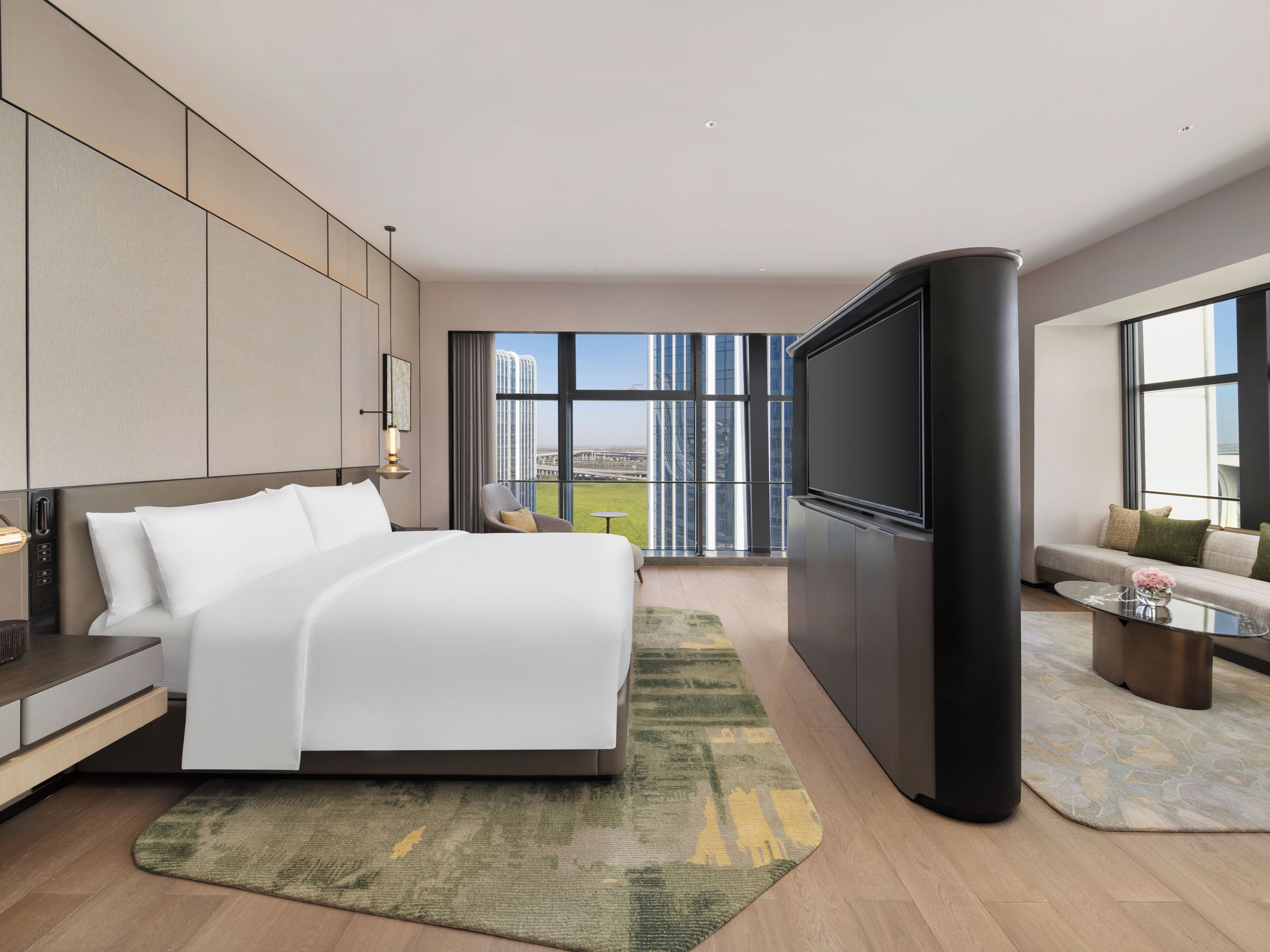 A modern hotel room with a large bed, a flat-screen TV, a sofa, and a view of the cityscape.