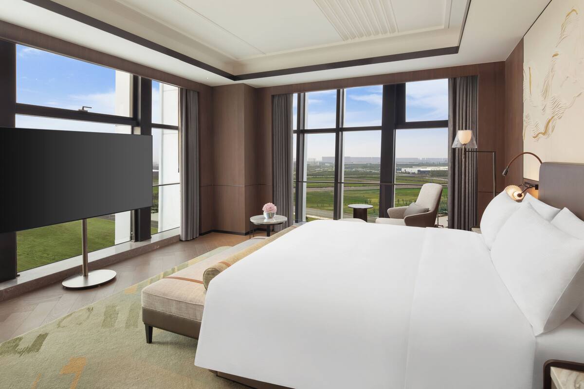 Ein Hotelzimmer mit einem großen Bett, einem Fernseher, einem Sessel, einem kleinen Tisch und Blick auf die Stadt.