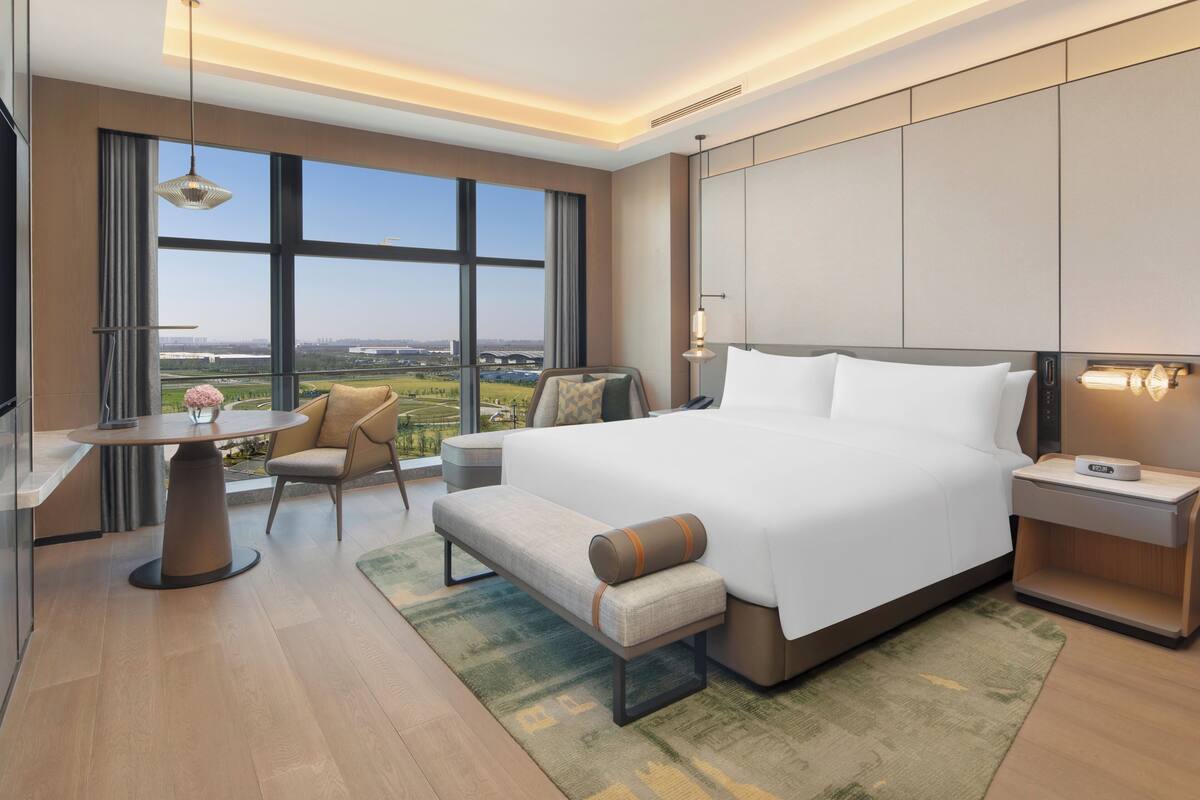 Ein modernes Hotelzimmer mit einem großen Bett, einem Schreibtisch, einem Sessel und Blick auf eine Stadtlandschaft.