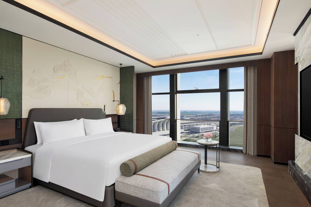 Ein modernes Hotelzimmer mit einem großen Bett, einem kleinen Tisch mit einer Vase, einem Fernseher und Blick auf die Stadt.