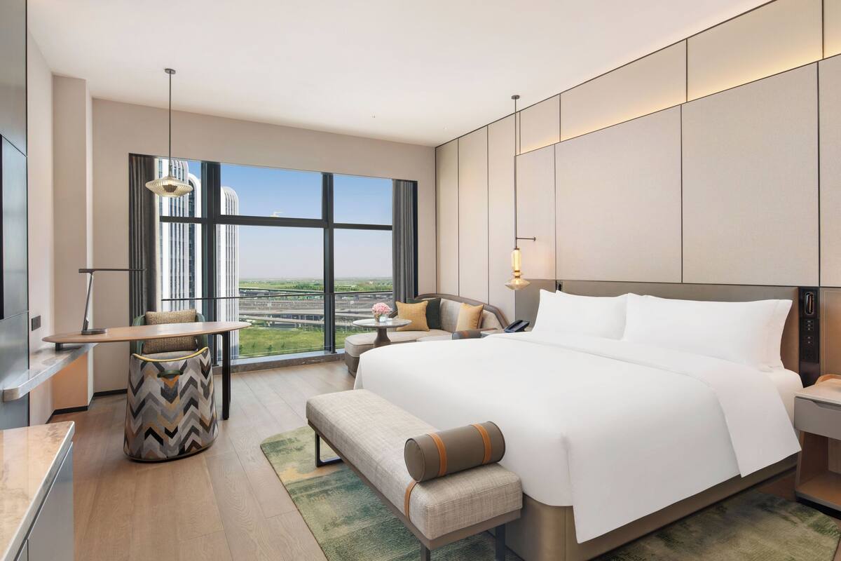 Ein modernes Hotelzimmer mit einem großen Bett, einem Schreibtisch, einem Sessel, einem kleinen Tisch und Blick auf die Stadt.
