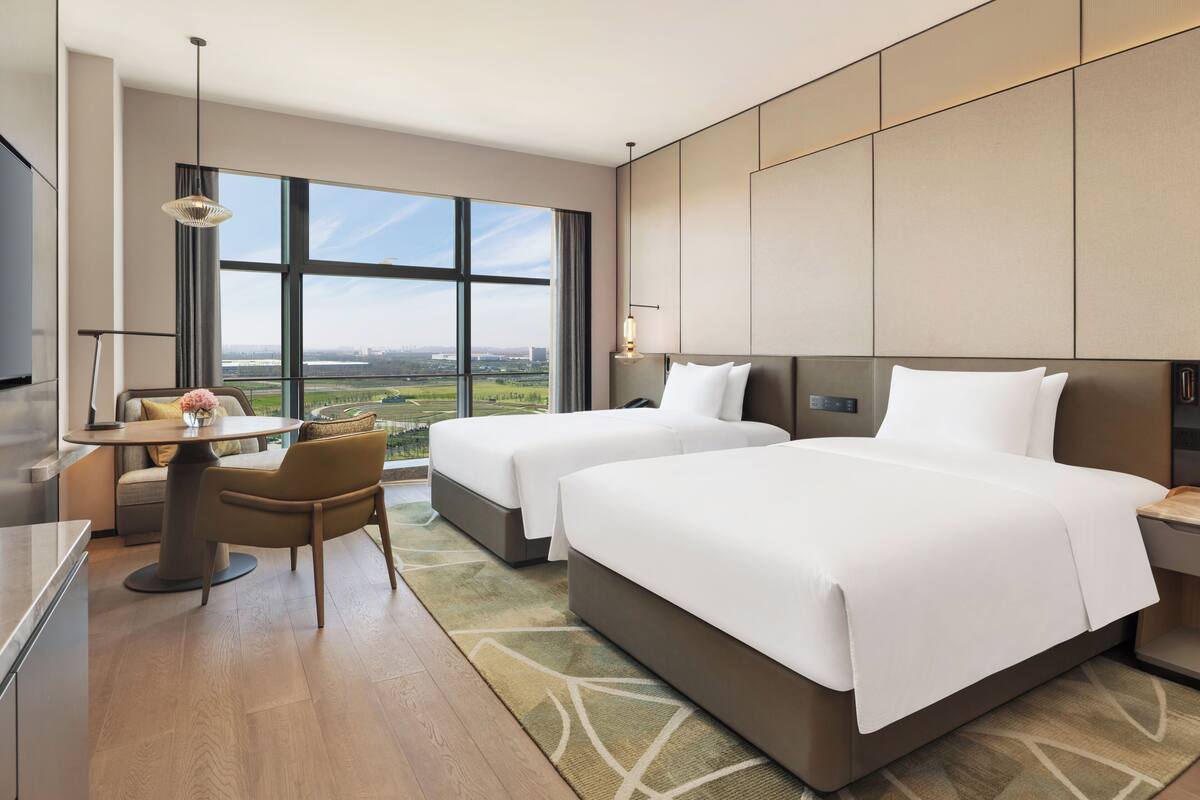 Ein Hotelzimmer mit zwei Betten, einem Schreibtisch, einem Sessel, einem Fernseher und großen Fenstern mit Blick auf die Stadt.