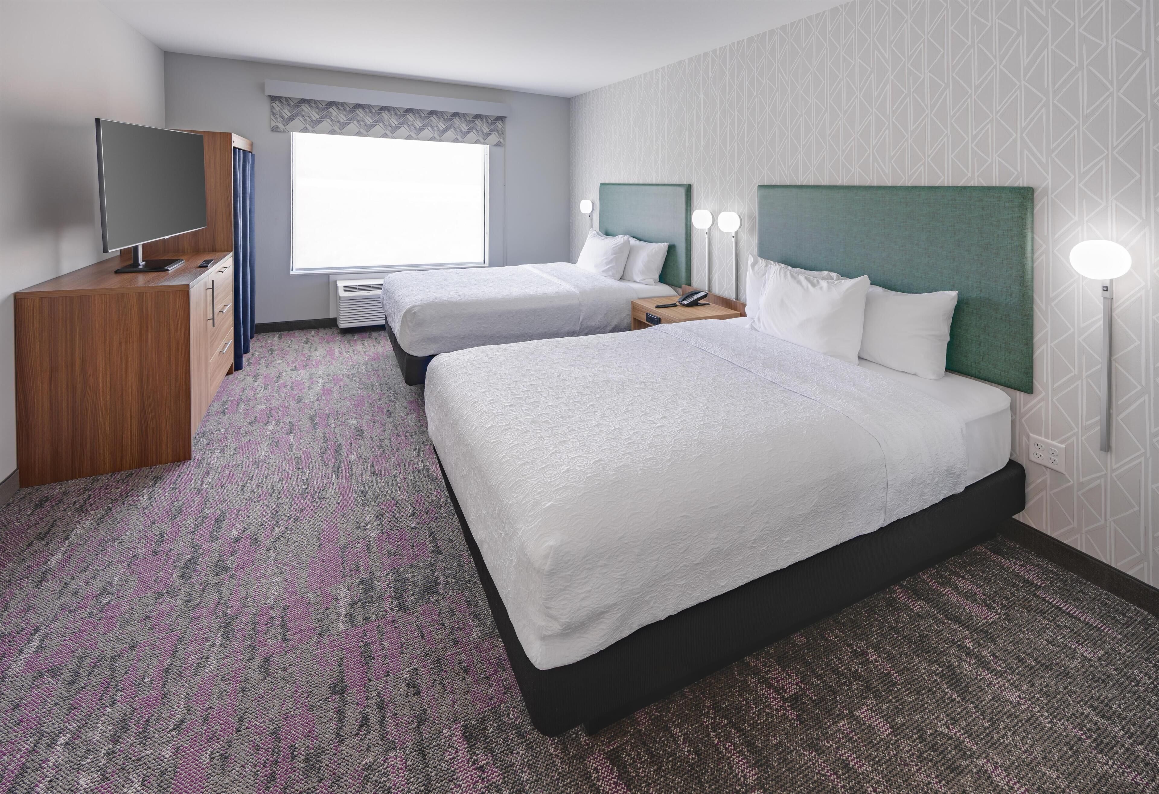 A hotel room with two beds, a TV, a desk, and a window with a view.