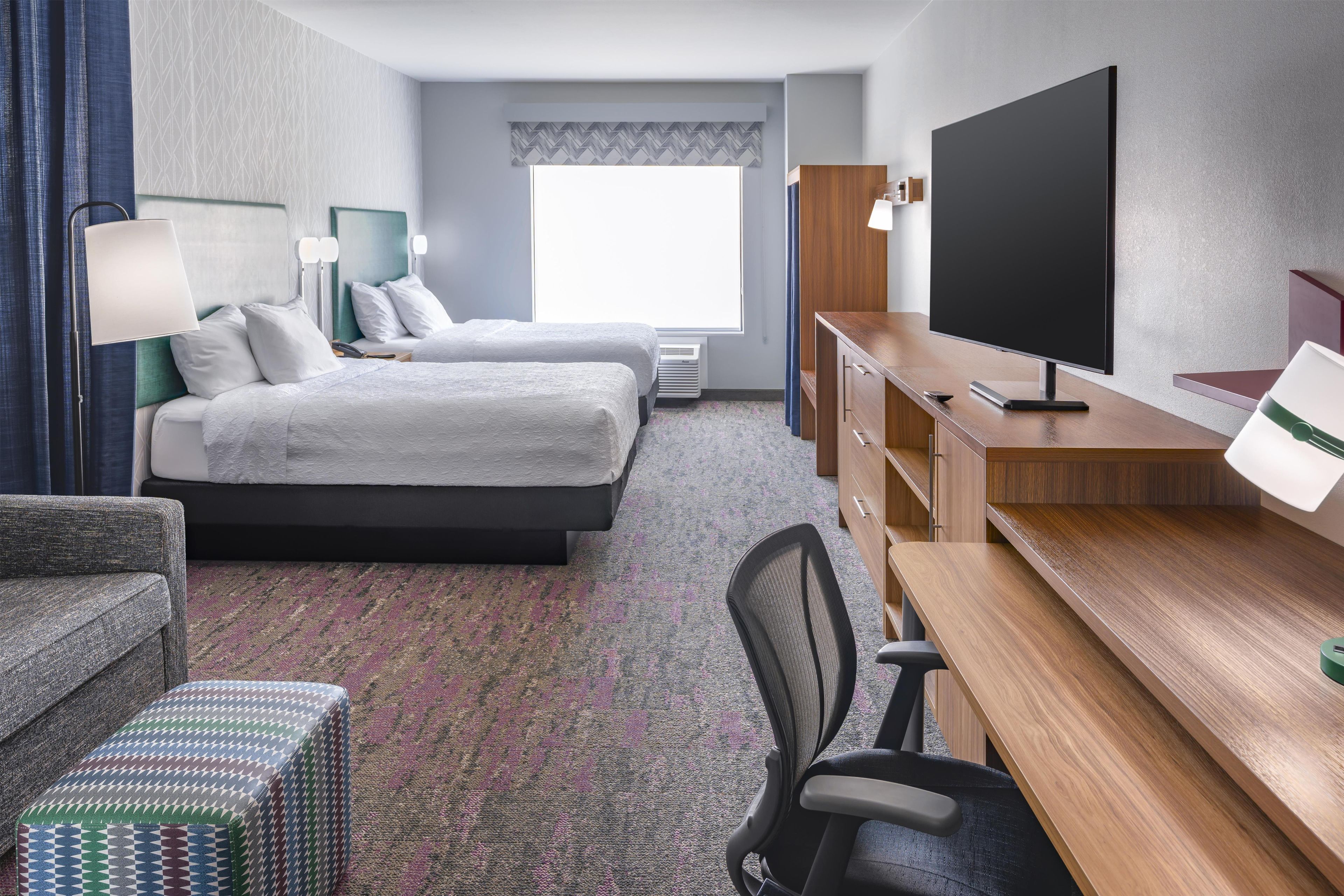 A hotel room with two beds, a desk with a flat-screen TV, a chair, a lamp, and a window with a view.