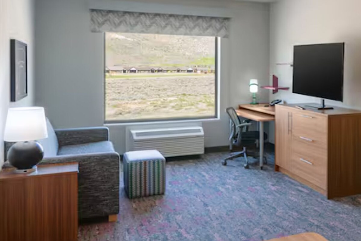 Habitación de hotel con escritorio, televisor, zona de comedor y ventana con vistas al exterior.