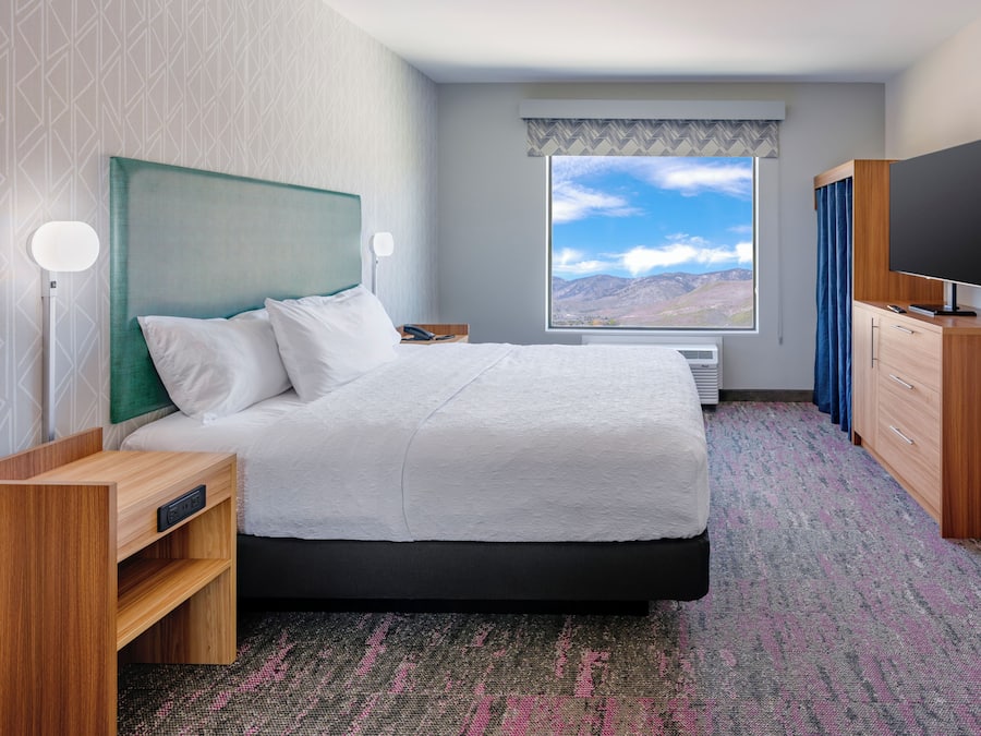 A hotel room with a bed, a TV, a desk, and a window with a mountain view.