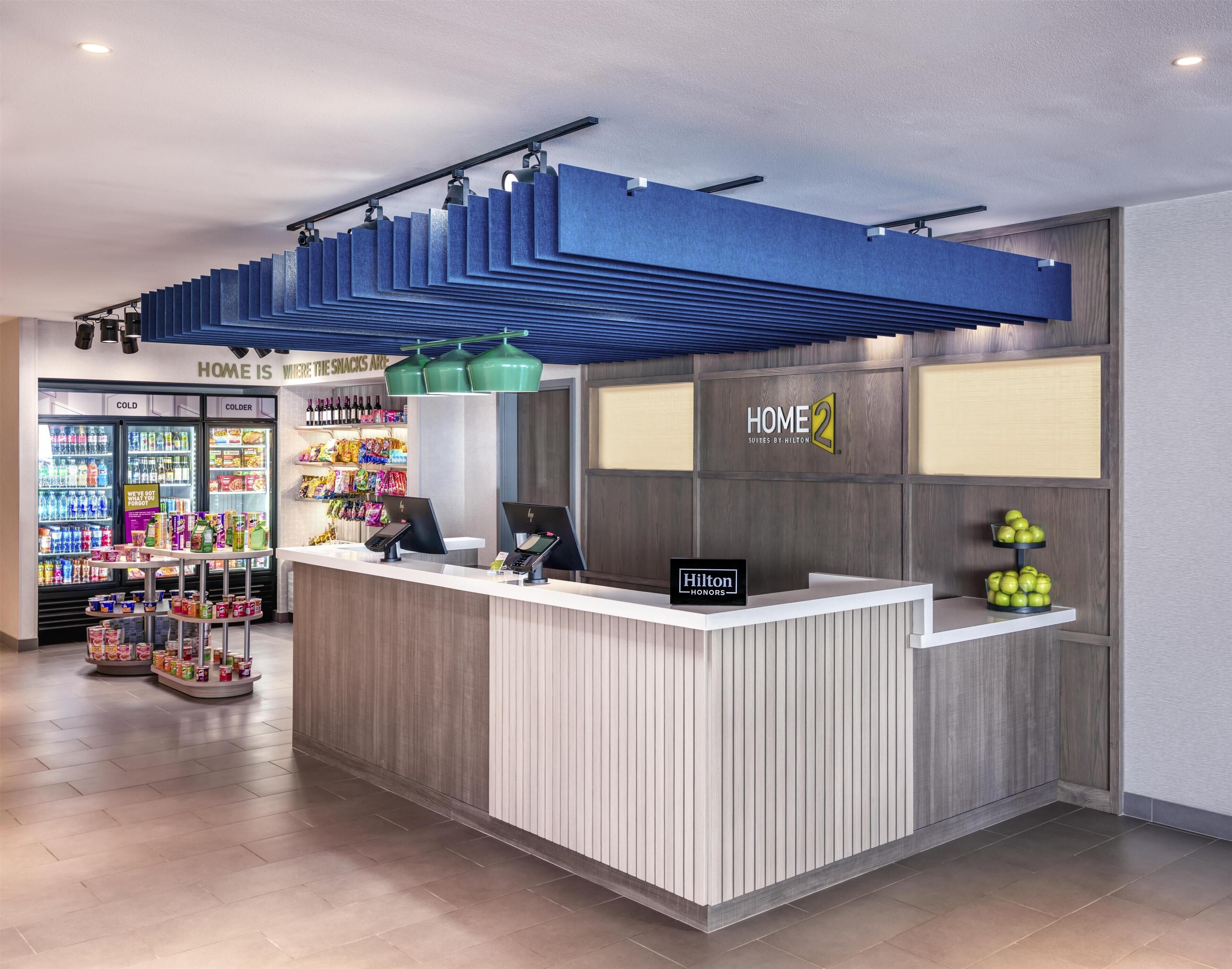 A hotel reception area with a counter, vending machines, and a sign for 'Home 2 Suites by Hilton.'